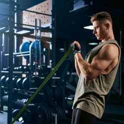 Portrait of muscular bodybuilder training arms with fitness resistance bands. Side view of man with perfect body training biceps in gym in dark atmosphere. Concept of bodybuilding.