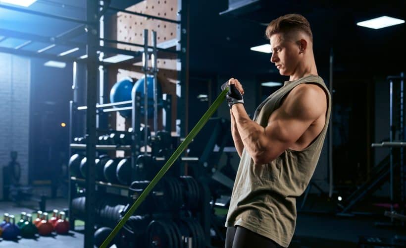 Portrait of muscular bodybuilder training arms with fitness resistance bands. Side view of man with perfect body training biceps in gym in dark atmosphere. Concept of bodybuilding.