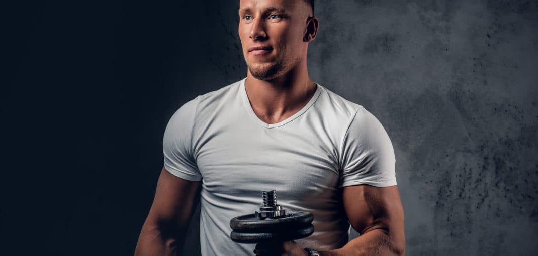 Handsome muscular blond male in a white t shirt holds dumbbell over grey artistic background.