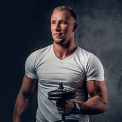 Handsome muscular blond male in a white t shirt holds dumbbell over grey artistic background.