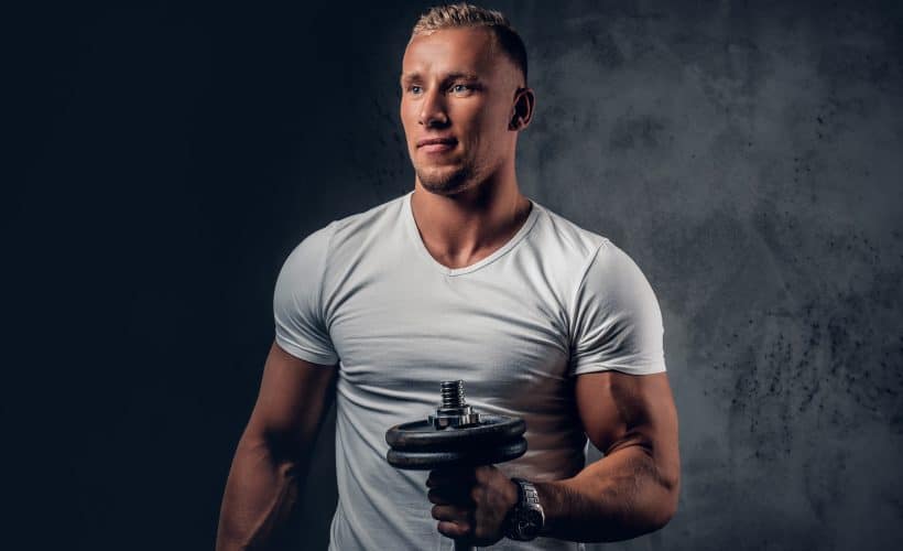 Handsome muscular blond male in a white t shirt holds dumbbell over grey artistic background.