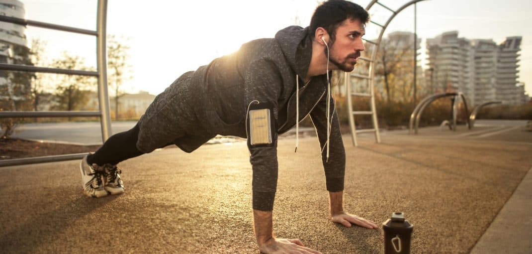 Man performing push-ups outdoors in a park during sunrise, wearing sportswear, earphones, and an armband for his phone.