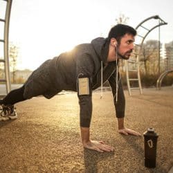 Man performing push-ups outdoors in a park during sunrise, wearing sportswear, earphones, and an armband for his phone.