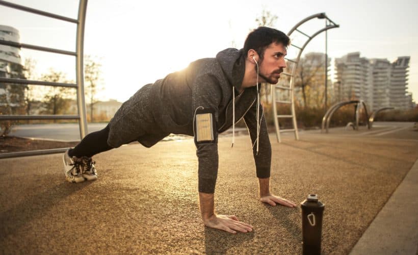 Man performing push-ups outdoors in a park during sunrise, wearing sportswear, earphones, and an armband for his phone.