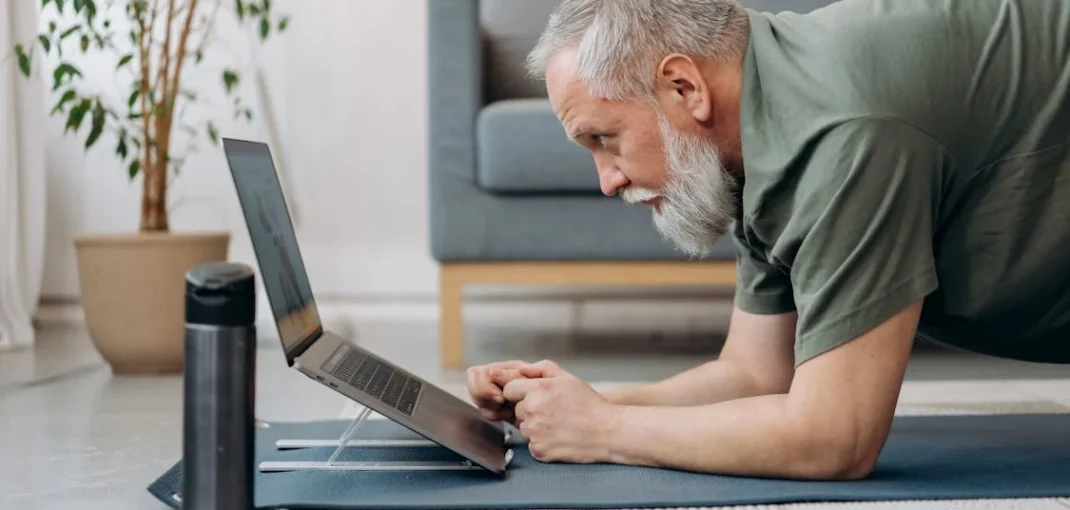 An older man lying on the floor in a plank while using a laptop.