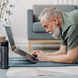 An older man lying on the floor in a plank while using a laptop.